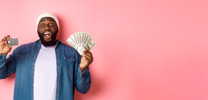 Shopping concept. Excited african-american man showing credit card and dollars, got deposit or money loan, standing over pink background.