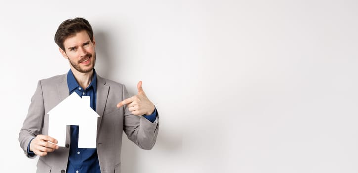 Real estate and insurance concept. Salesman in grey suit showing paper house cutout, selling property, smiling at camera, white background.