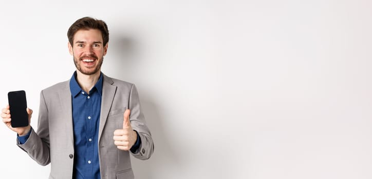 E-commerce and online shopping concept. Satisfied businessman in suit showing empty smartphone screen and thumb-up, praise good deal, white background.