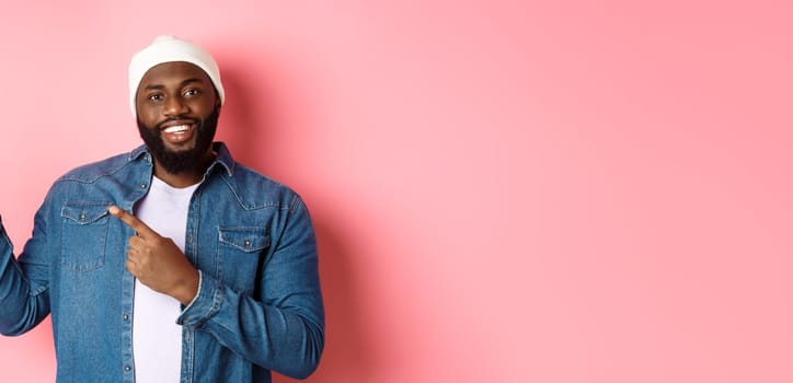 Happy african american man pointing fingers upper left corner, showing promo offer logo, smiling pleased, wearing beanie with denim jacket, pink background.