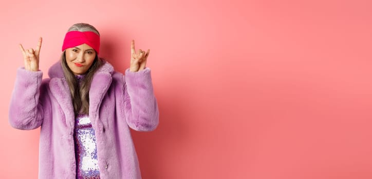 Cool asian mature woman in fashionable faux fur coat, showing rock-n-roll horns signs and smiling sassy at camera, having fun, standing over pink background.