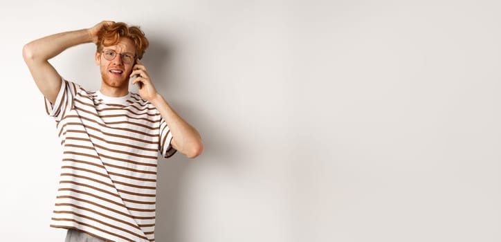 Puzzled redhead guy talking on phone, scratching head and looking confused or indecisive, standing over white background.