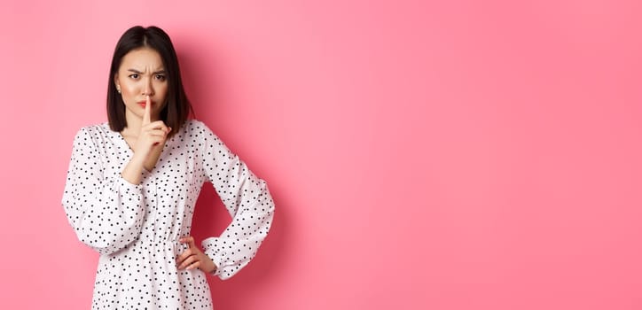 Angry asian female model shushing, frowning and hushing, showing taboo gesture, standing displeased against pink background, demand silence.