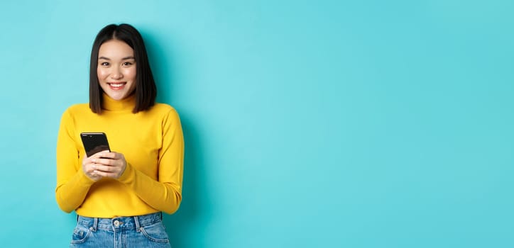 Beautiful and stylish asian woman shopping online on mobile phone, standing over blue background.