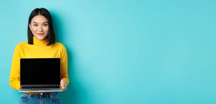 Beautiful and stylish asian woman demonstrate product on screen, showing empty laptop display and smiling, standing over blue background.