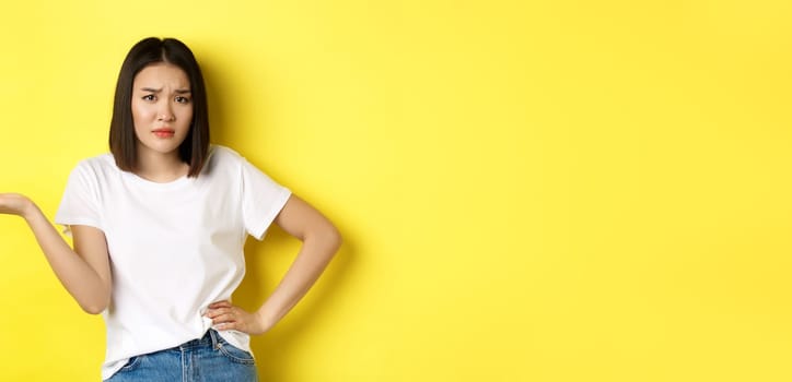 Disappointed asian woman asking so what, raise hand up and staring skeptical at camera, standing over yellow background.