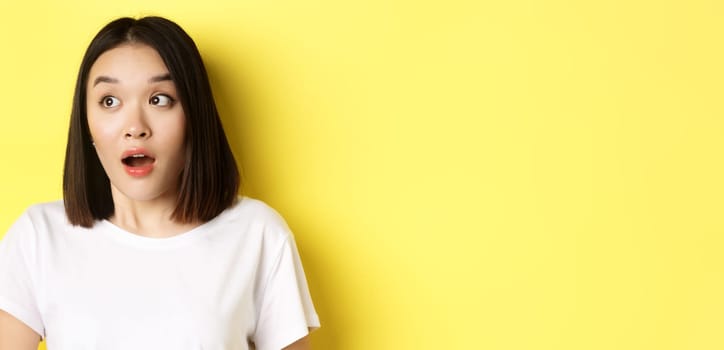 Close up of surprised asian girl drop jaw, gasping and looking left at logo, standing over yellow background.