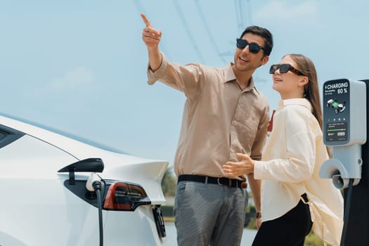 Young couple recharge electric car's battery from charging station in outdoor green city park in springtime. Rechargeable EV car for sustainable environmental friendly urban travel lifestyle.Expedient