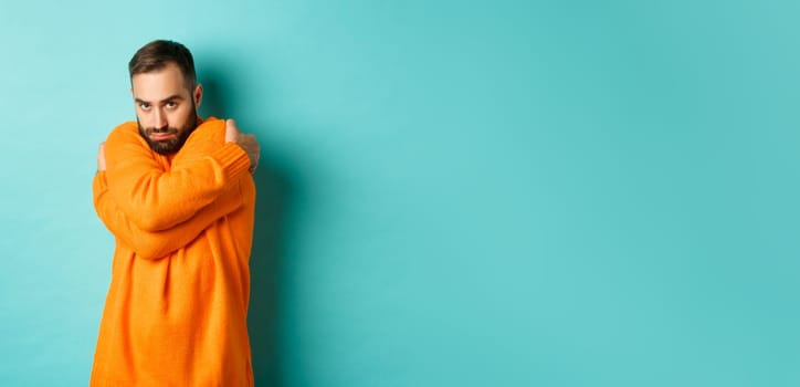 Timid man feeling offended and defensive, hugging himself and looking suspicious at camera, standing over light blue background.