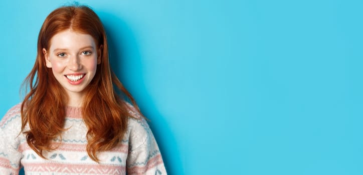 Close-up of cute redhead girl in sweater smiling happy at camera, standing against blue background.