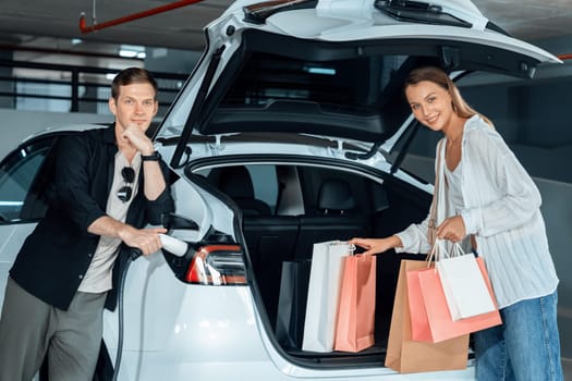 Young couple travel with EV electric car to shopping center parking lot charging in downtown city showing urban sustainability lifestyle by green clean rechargeable energy of electric vehicle innards