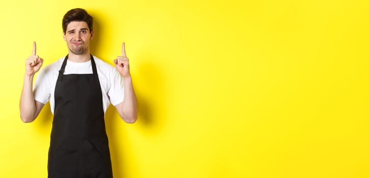 Displeased and skeptical waiter in black apron pointing fingers up, express dislike and indifference, standing over yellow background.