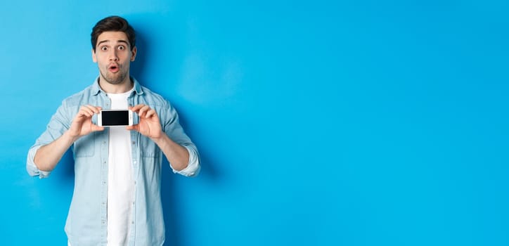 Surprised guy showing mobile phone screen and looking impressed, standing over blue background.