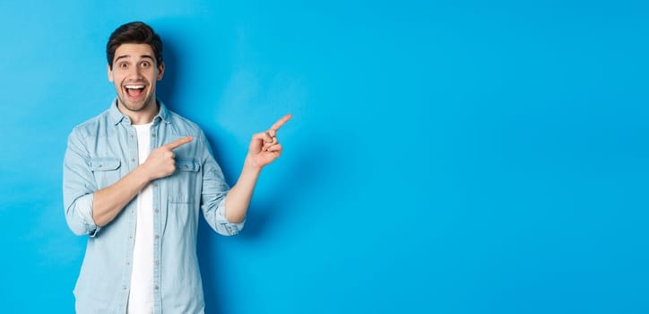 Image of excited handsome man in casual outfit, showing advertisement, pointing fingers right at copy space and smiling, standing against blue background.