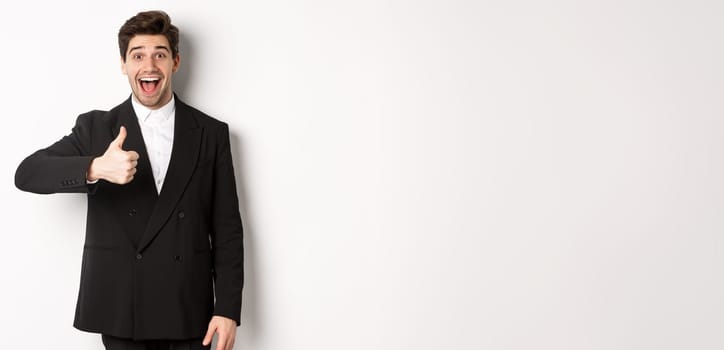 Portrait of amazed and impressed handsome man in black suit, showing thumb-up, praise awesome product, liking something good, standing against white background.