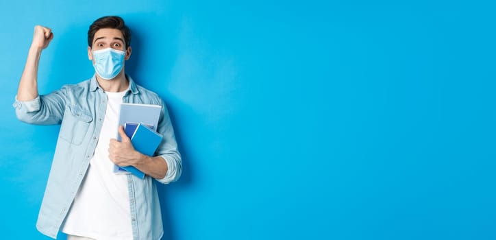 Education, covid-19 and social distancing. Excited male student in medical mask triumphing, raising fist up and holding notebooks, standing over blue background.