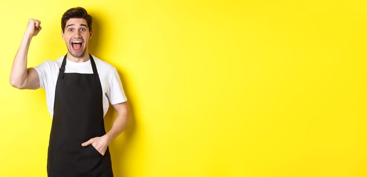 Cheerful seller making fist pump, rejoicing and triumphing, standing in black apron against yellow background.