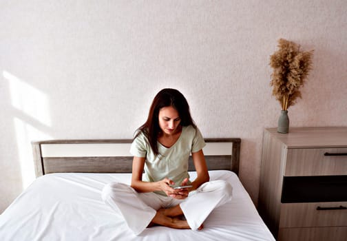 Brunette woman sitting in white bed using smartphone, having video call, smiling young woman relaxing in bedroom, waking up in the morning and sending text messages on one device, technology concept.