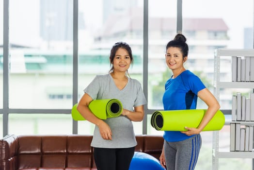 Asian adult and young woman smiling holding a yoga mat after yoga and exercise. Portrait of happy beautiful female standing hold yoga mat indoor studio, sport healthy workout concept