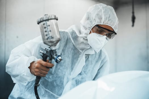 Automotive service worker in full protective gear expertly apply color paint in to car's bodywork with spray gun or respirator painting in chamber workshop. Car paint service for scratch refinish.Oxus