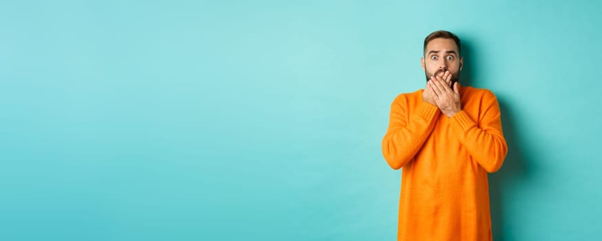 Startled man looking at horrible scene, gasping and stare scared, standing in orange winter sweater, studio background.