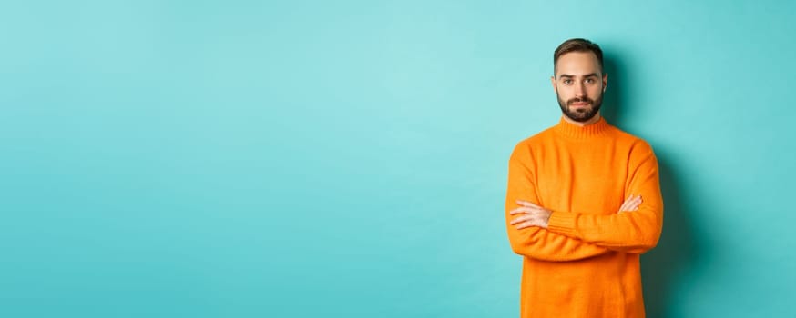 Confident young man looking determined, cross arms on chest, wearing orange winter sweater, standing against turquoise studio background.
