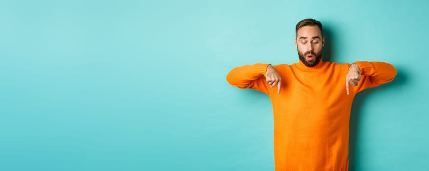 Amazed guy in orange sweater, pointing fingers down and looking with interest at promo, holiday discounts, standing over turquoise background.