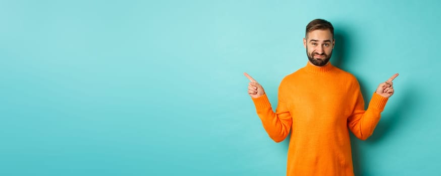 Doubtful and indecisive man pointing fingers sideways, showing different sides, two choices, looking skeptical, standing over light blue background.