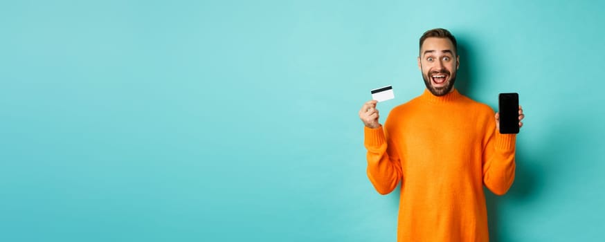 Online shopping. Excited man showing mobile screen and credit card, smiling amazed, standing over turquoise background.
