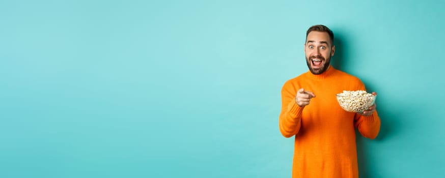 Handsome young man holding bowl of popcorn, smiling amazed and pointing at camera, standing over blue background.