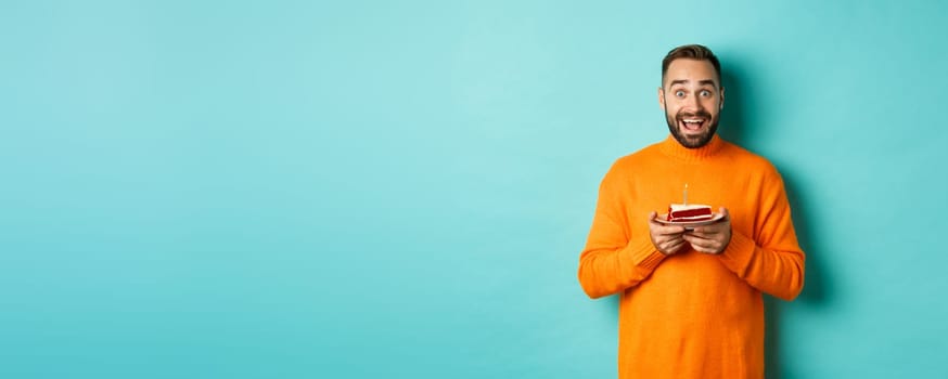 Happy adult man celebrating birthday, holding bday cake with candle and smiling, standing against turquoise background.