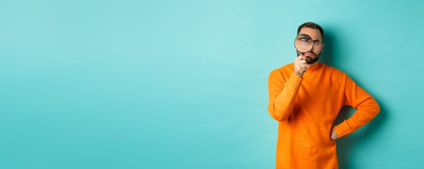 Funny man looking serious through magnifying glass, inspecting something, standing in orange sweater against turquoise background.