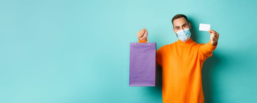 Covid-19, pandemic and lifestyle concept. Happy male customer in face mask showing credit card and purple shopping bag, standing over light blue background.