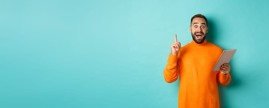 Excited adult man using digital tablet and having an idea, raising finger eureka sign, standing over light blue background.