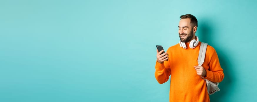 Caucasian man with headphones and backpack looking at phone, reading message and smiling, standing over turquoise background.