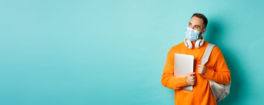 Handsome caucasian man with headphones and backpack, holding laptop and wearing medical mask, looking happy, standing over light blue background.