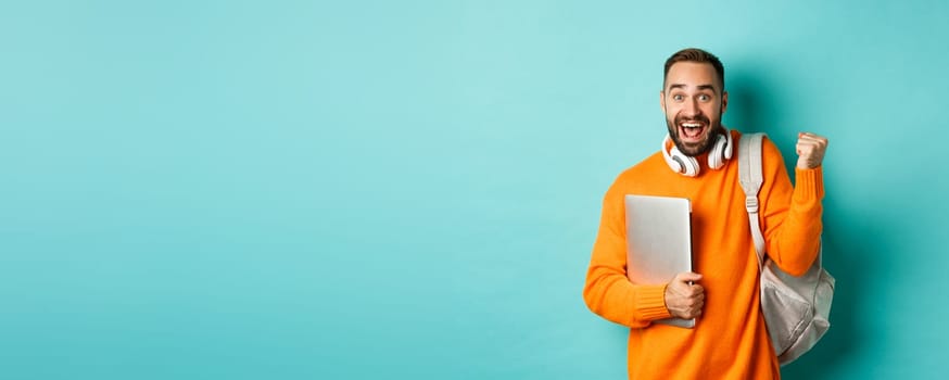 Happy man with backpack and headphones, holding laptop and smiling, cheering of win, triumphing over turquoise background.