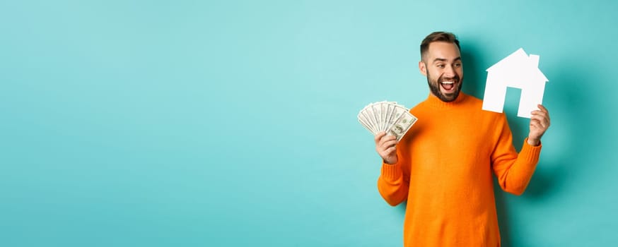 Real estate concept and mortgage concept. Cheerful man holding money and paper house, smiling excited, buying or renting flat, standing over blue background.