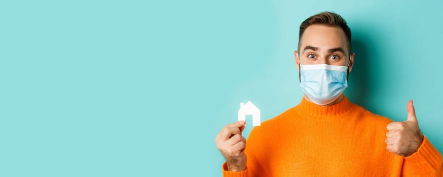 Real estate and coronavirus pandemic concept. Close-up of adult man in medical mask holding small paper house maket and smiling, showng thumb-up in approval.