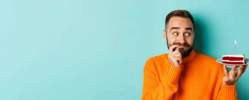 Close-up of happy adult man celebrating birthday, holding bday cake with candle and making wish, standing against turquoise background.