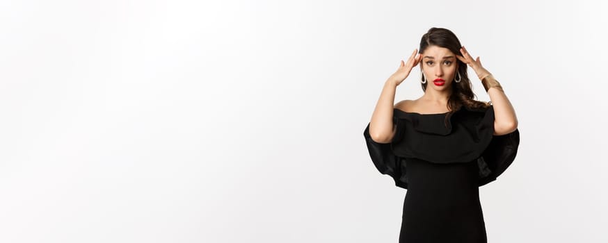 Fashion and beauty. Annoyed and tired woman in black dress, touching head and roll eyes bothered, standing distressed against white background.