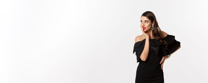 Fashion and beauty. Image of stylish beautiful woman in black dress and makeup, looking left with temptation, touching red lips, standing over white background.