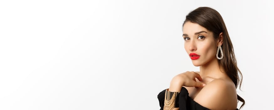 Fashion and beauty concept. Close-up of elegant woman with red lips, makeup and earrings, looking at camera self-assured, standing over white background.