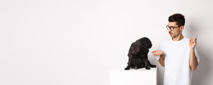 Handsome young man teaching dog commands, talking to cute black pug, standing over white background.