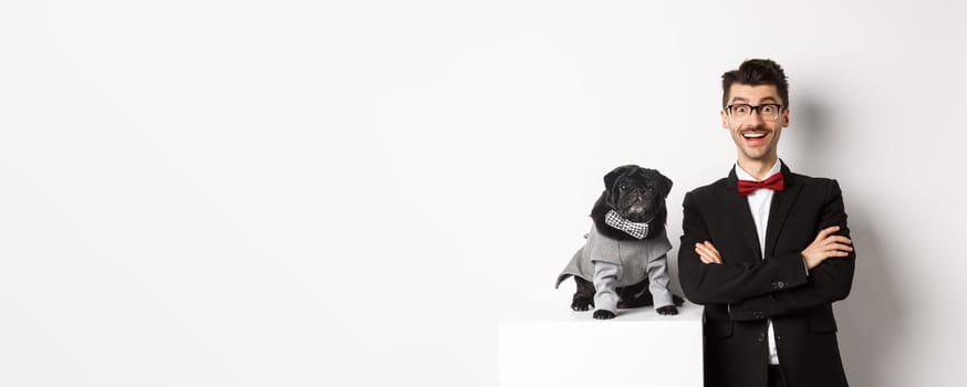 Animals, party and celebration concept. Happy dog owner in suit and puppy in costume looking excited at camera, having fun, standing over white background.