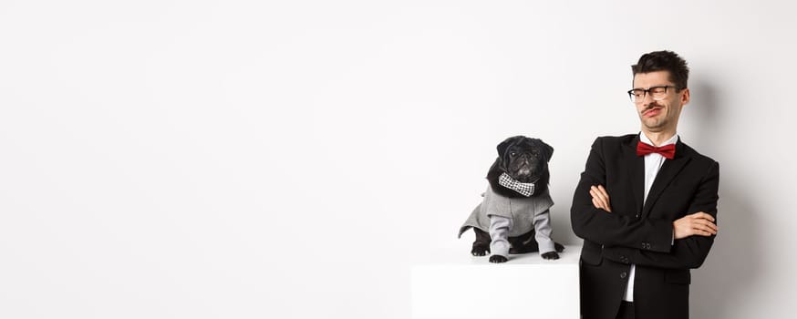 Animals, party and celebration concept. Image of funny young man in suit and glasses, looking skeptical at cute pug in costume, standing over white background.
