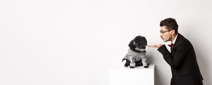 Pets, winter holidays and celebration concept. Handsome young man sending air kiss at cute black puppy wearing costume for New Year, owner standing in suit over white background.