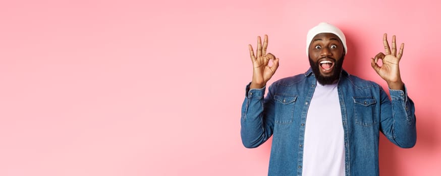 Impressed Black man in beanie and shirt, showing ok signs and staring at camera amazed, smiling happy, praise great offer, recommending product, pink background.