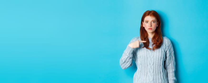 Nervous and confused redhead girl pointing at herself, standing in sweater against blue background.