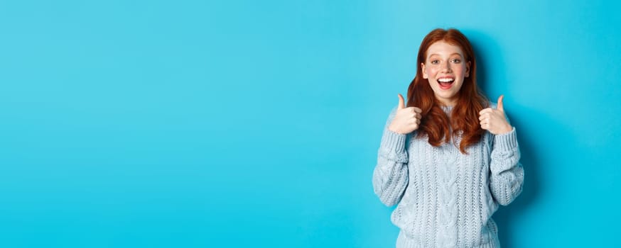 Supportive redhead girl in sweater, showing thumbs-up and looking amazed, praising good choice, standing over blue background.